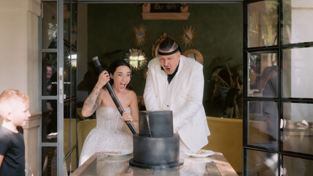 Morrhof Hochzeit Ein Paar bereitet sich freudig darauf vor, eine zweistöckige schwarze Torte anzuschneiden. Zu sehen ist eine Frau in einem glitzernden schulterfreien Kleid und ein Mann im weißen Anzug der aufgeregt mit einem Schwert die Hochzeitstorte zerteilt