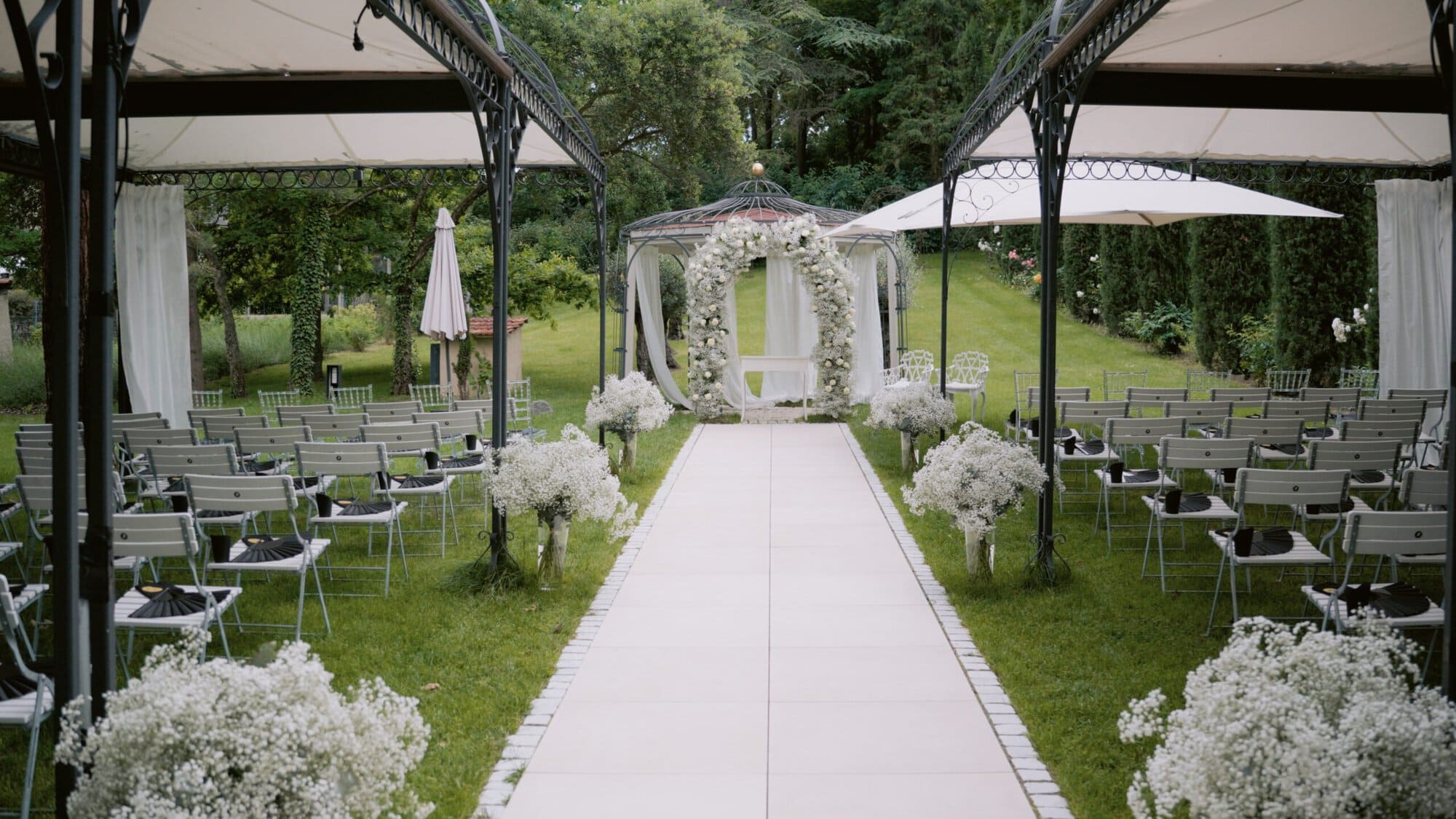 Morrhof Hochzeit Hochzeitsaufbau im Freien mit einem weißen Gang, der zu einem Blumenbogen führt. Stuhlreihen säumen beide Seiten des Ganges, und weiße Blumen schmücken den Weg. Zwei Baldachine spenden Schatten, umgeben von üppigem Grün.