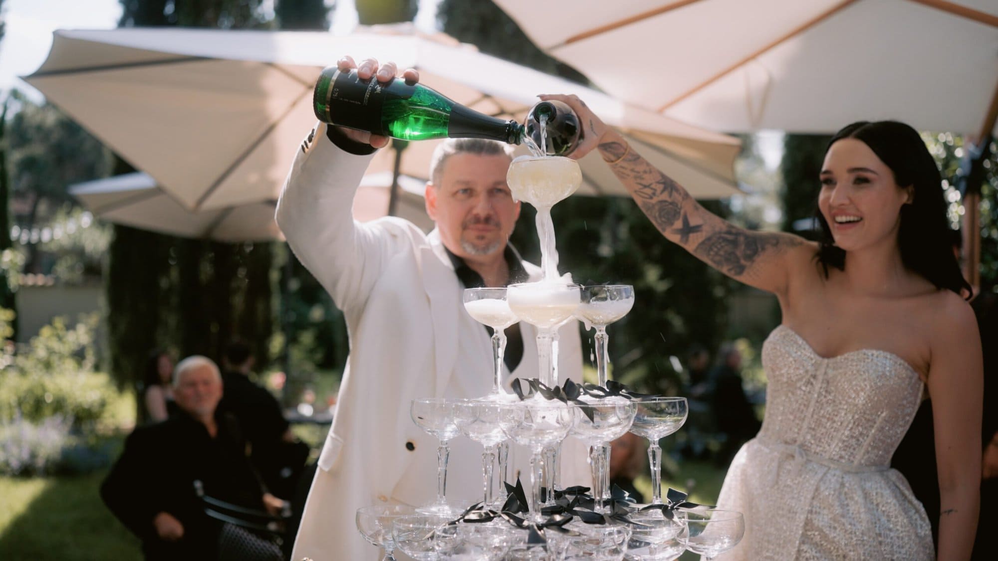 Morrhof Hochzeit Braut und Bräutigam gießen bei einer Hochzeitsfeier im Freien Champagner in eine Pyramide aus Gläsern. Der Bräutigam trägt einen weißen Anzug und die Braut ein trägerloses weißes Kleid. Sie sind von Bäumen und Sonnenschirmen umgeben.