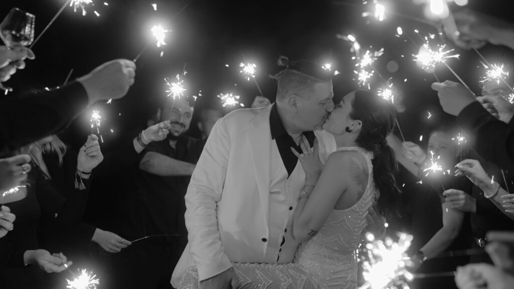 Morrhof Hochzeit Ein Paar küsst sich, umgeben von einer Gruppe von Menschen mit Wunderkerzen, was eine festliche, lichterfüllte Atmosphäre schafft. Der Mann trägt einen weißen Anzug und die Frau ein Paillettenkleid. Die Szene vermittelt Feierlichkeit und Freude.