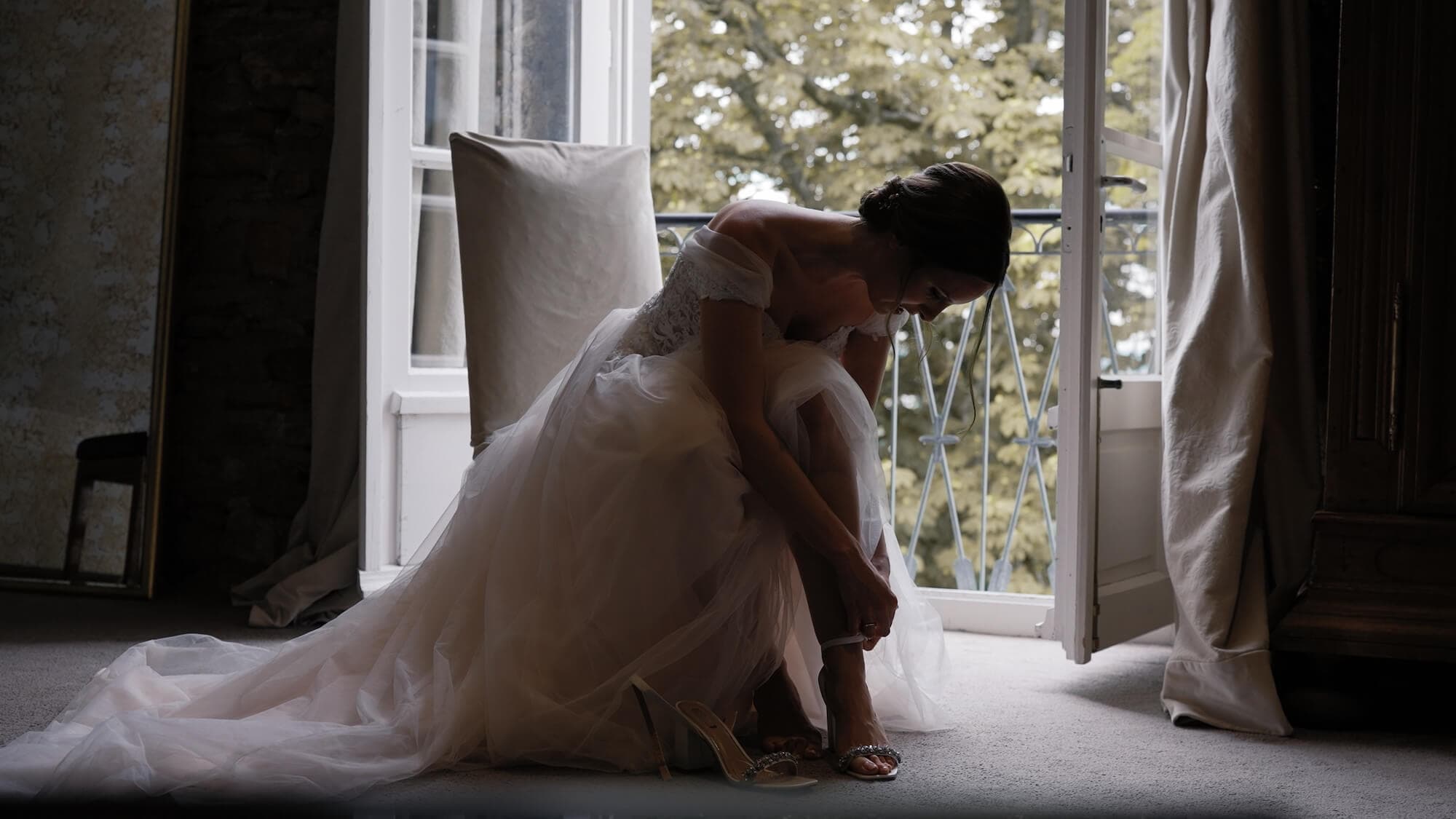 Hochzeitsvideo München - Hochzeitsvideograf Braut sitzt in der Getting ready Suite neben einer offenen Balkontür und richtet ihren Schuh.