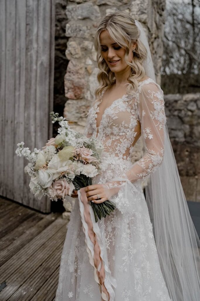 Hochzeitsvideo München - Hochzeitsvideograf Braut in einem Brautkleid aus floraler Spitze hält einen Blumenstrauß und steht neben einer Steinmauer.